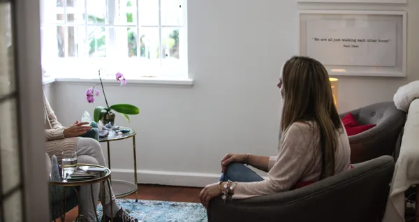Two people talking in a bright therapy room, one seated in a grey armchair