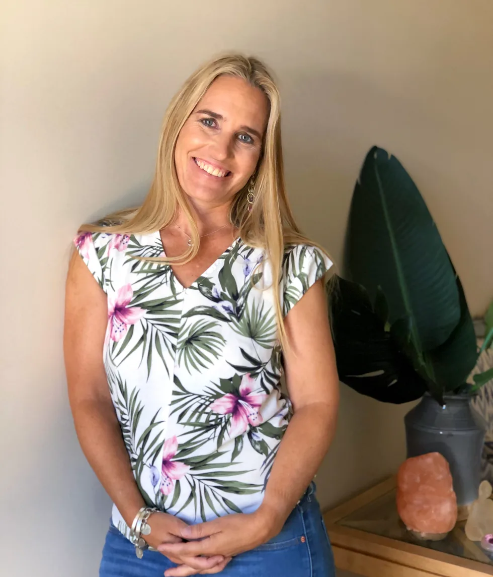 Renée Shearing smiling, in a floral top, beside a large green plant