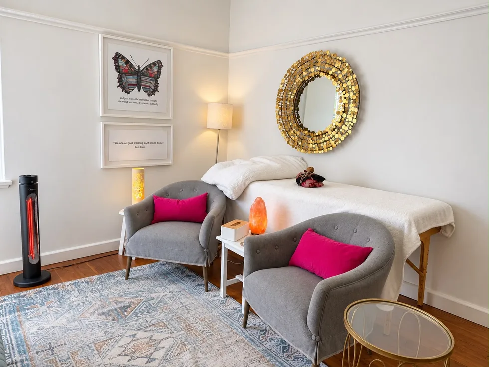 Two grey armchairs with pink cushions in Renée's consulting room, beside a treatment table, under a round gold mirror and a butterfly print