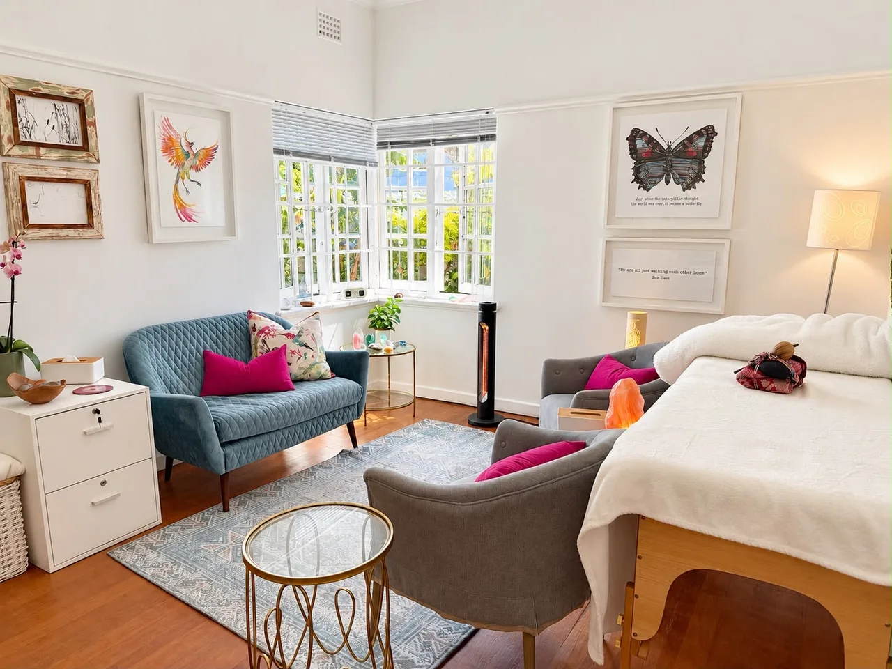 Renée’s consulting room in Wynberg: a teal sofa by the corner windows, two grey armchairs with pink cushions, a treatment table on the right, a patterned rug, and her phoenix painting and a butterfly print on the walls